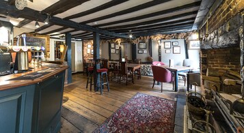 A rustic pub with exposed wooden beams, a bar area with stools, and a traditional fireplace.