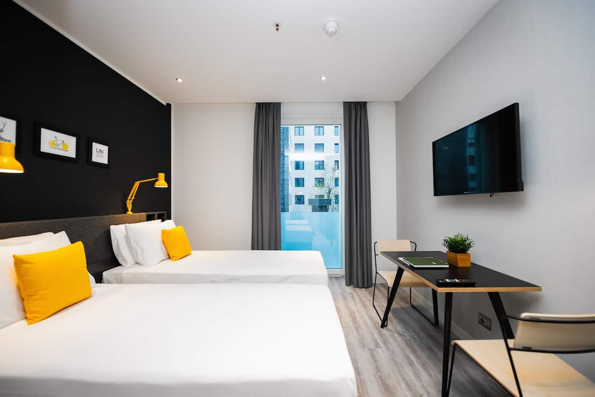 A hotel room with two beds, a desk, a TV, and a view of a building through the window.