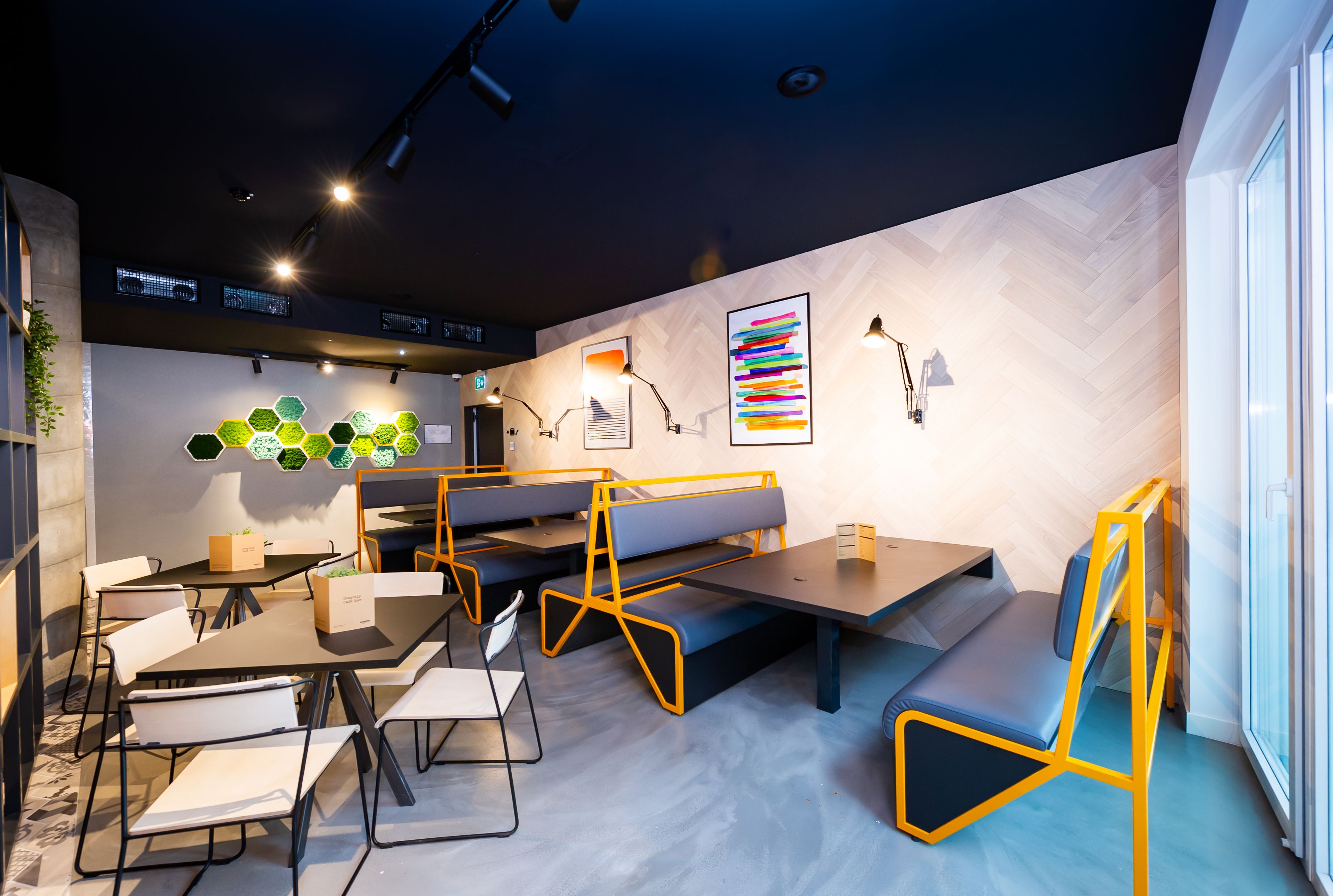 Un comedor moderno con mesas, bancos y sillas, que destaca por un techo de madera en espiga y un amplio ventanal a la derecha.