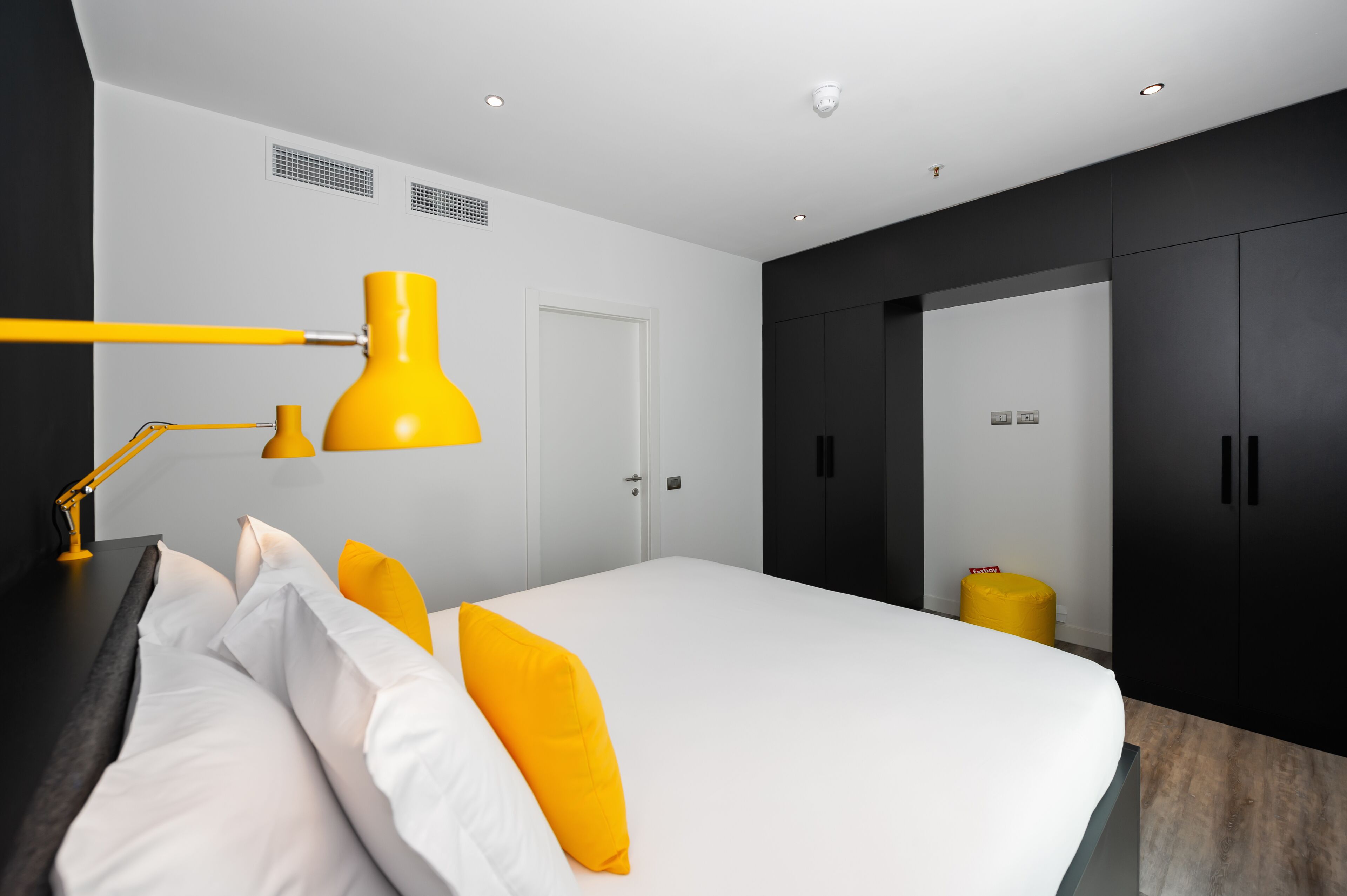 Un dormitorio moderno con una cama, un escritorio, una lámpara de escritorio amarilla y un armario.