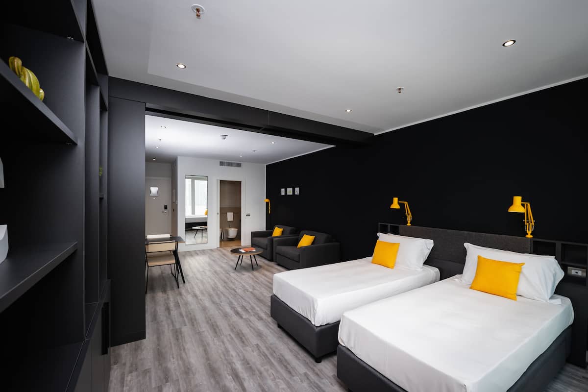 A modern hotel room with two beds, a desk, and a sofa.