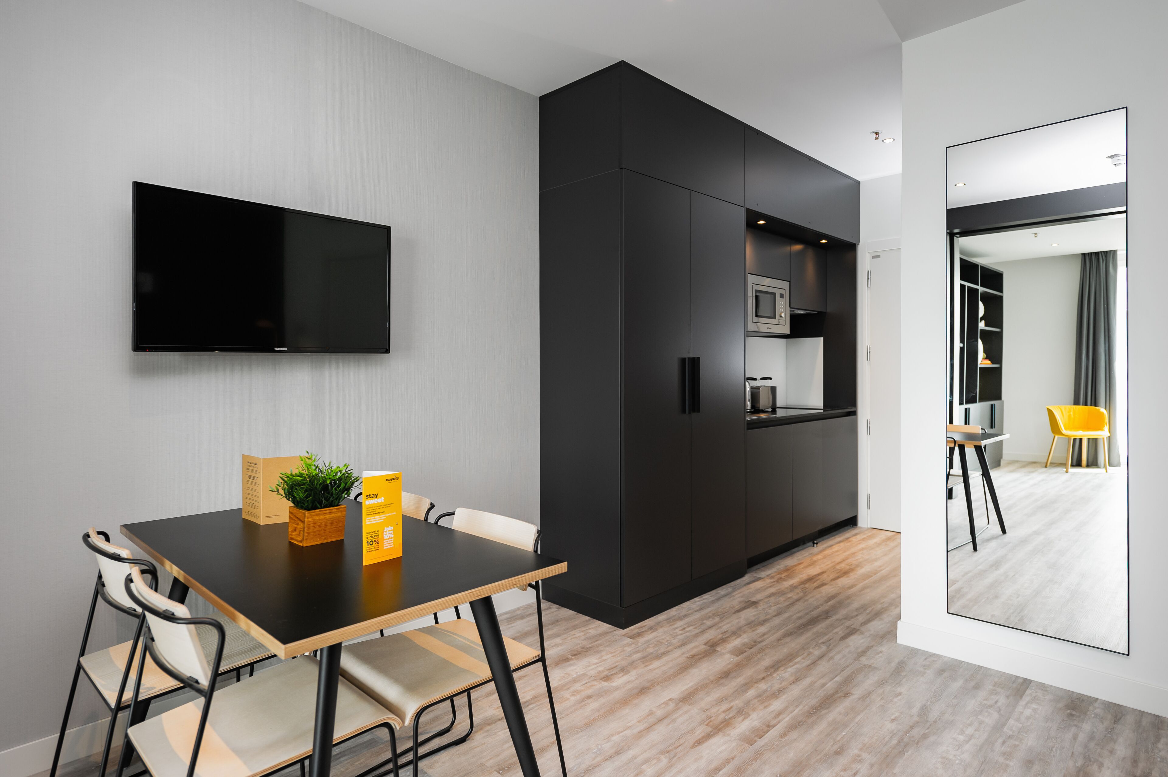 A modern kitchen with a flat-screen TV, a dining table with chairs, a small plant, and a yellow box.