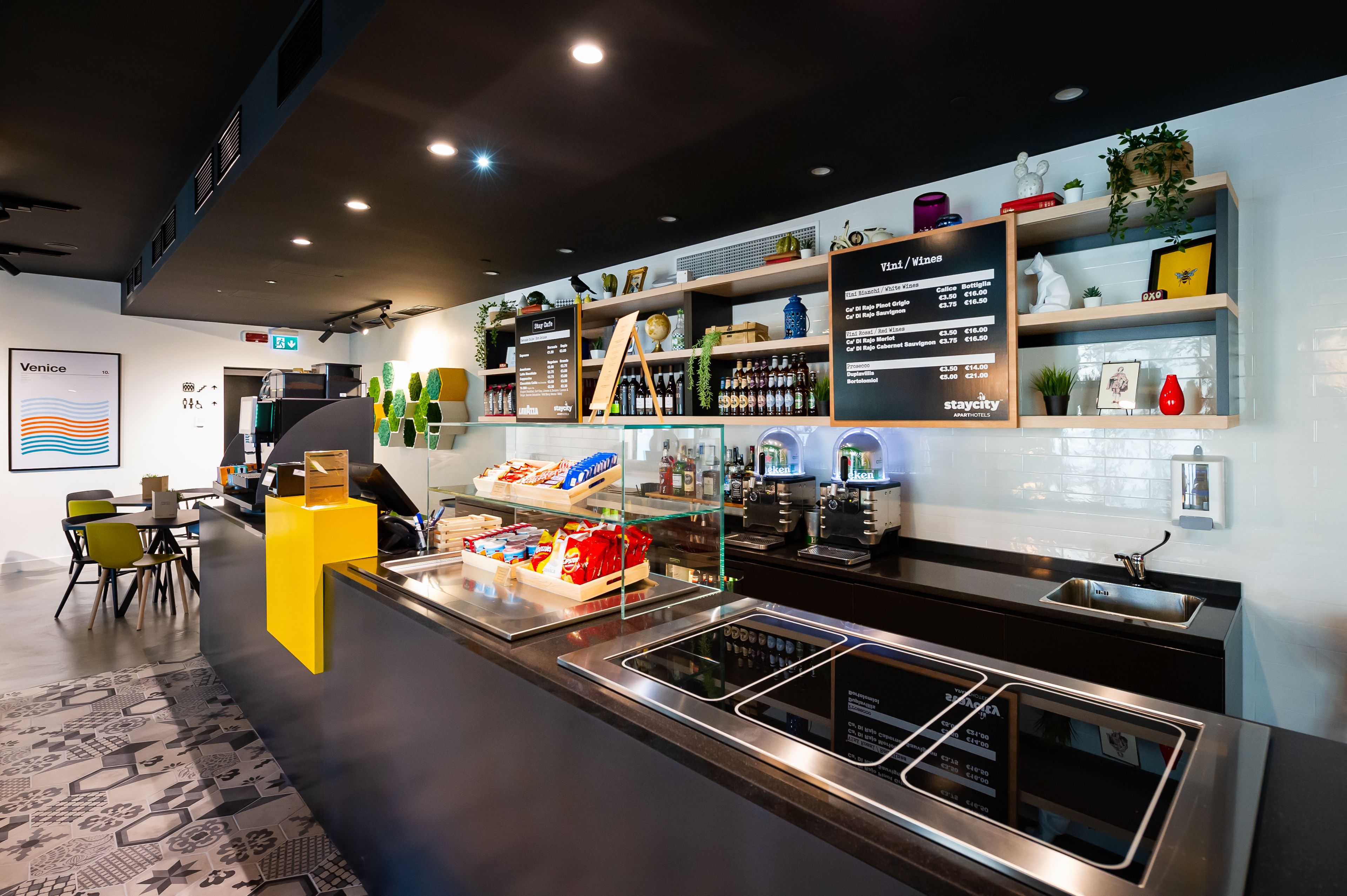 A modern bar area with a menu board, shelves with bottles, and a seating area with chairs.
