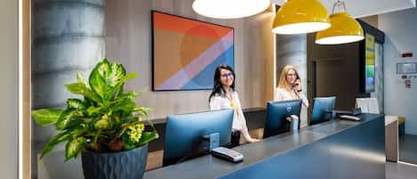 A reception desk with two employees, a large potted plant, and a colorful abstract painting on the wall.