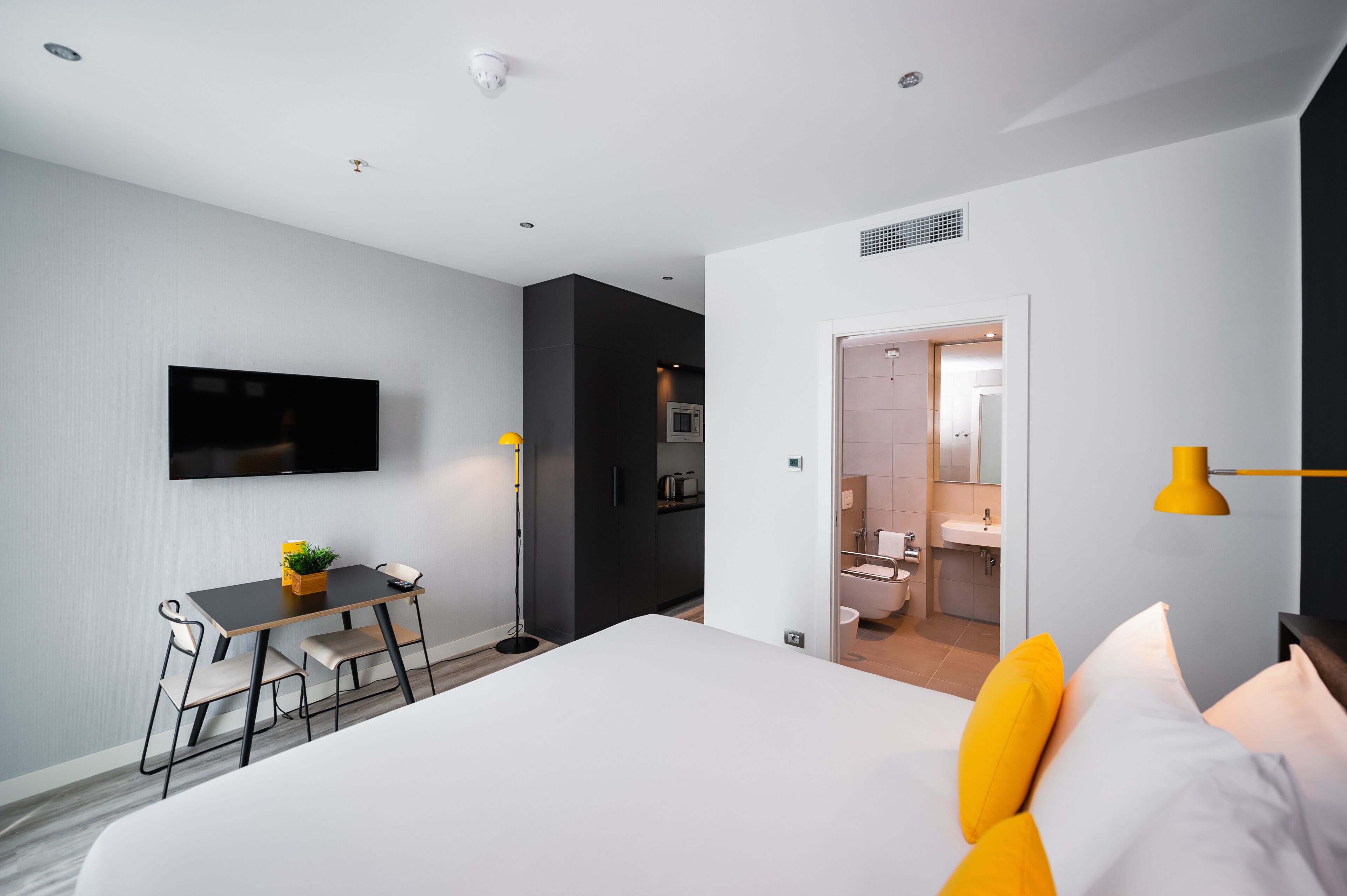 A modern hotel room with a bed, a desk, a flat-screen TV, a small table with a plant, and a bathroom visible through an open door.