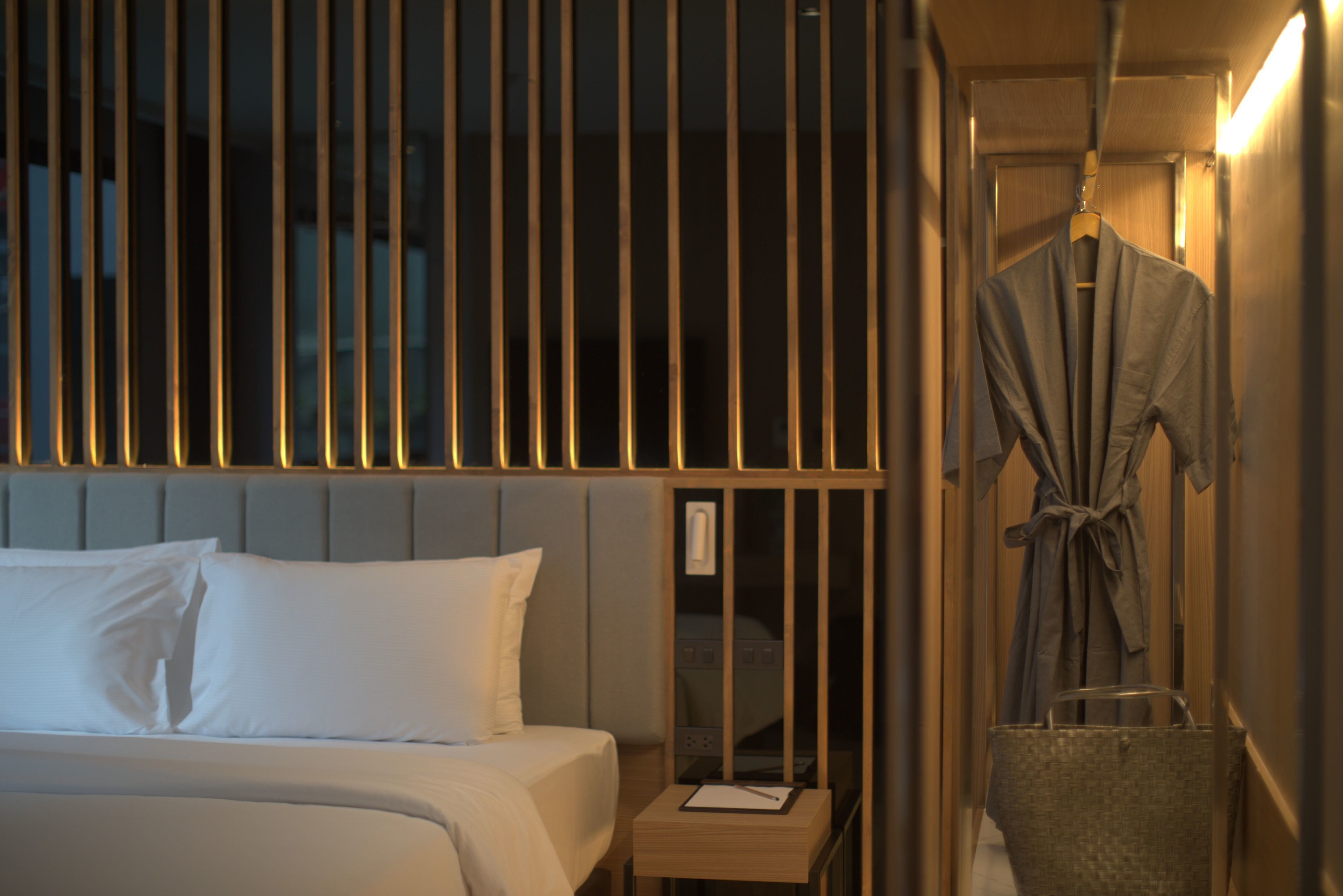 A hotel room with a bed, a bedside table with a lamp, a robe hanging on the wall, and a suitcase on the floor.