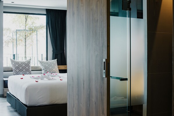 Quarto de hotel moderno com uma cama grande, box de vidro para o chuveiro e vista para uma árvore na janela.