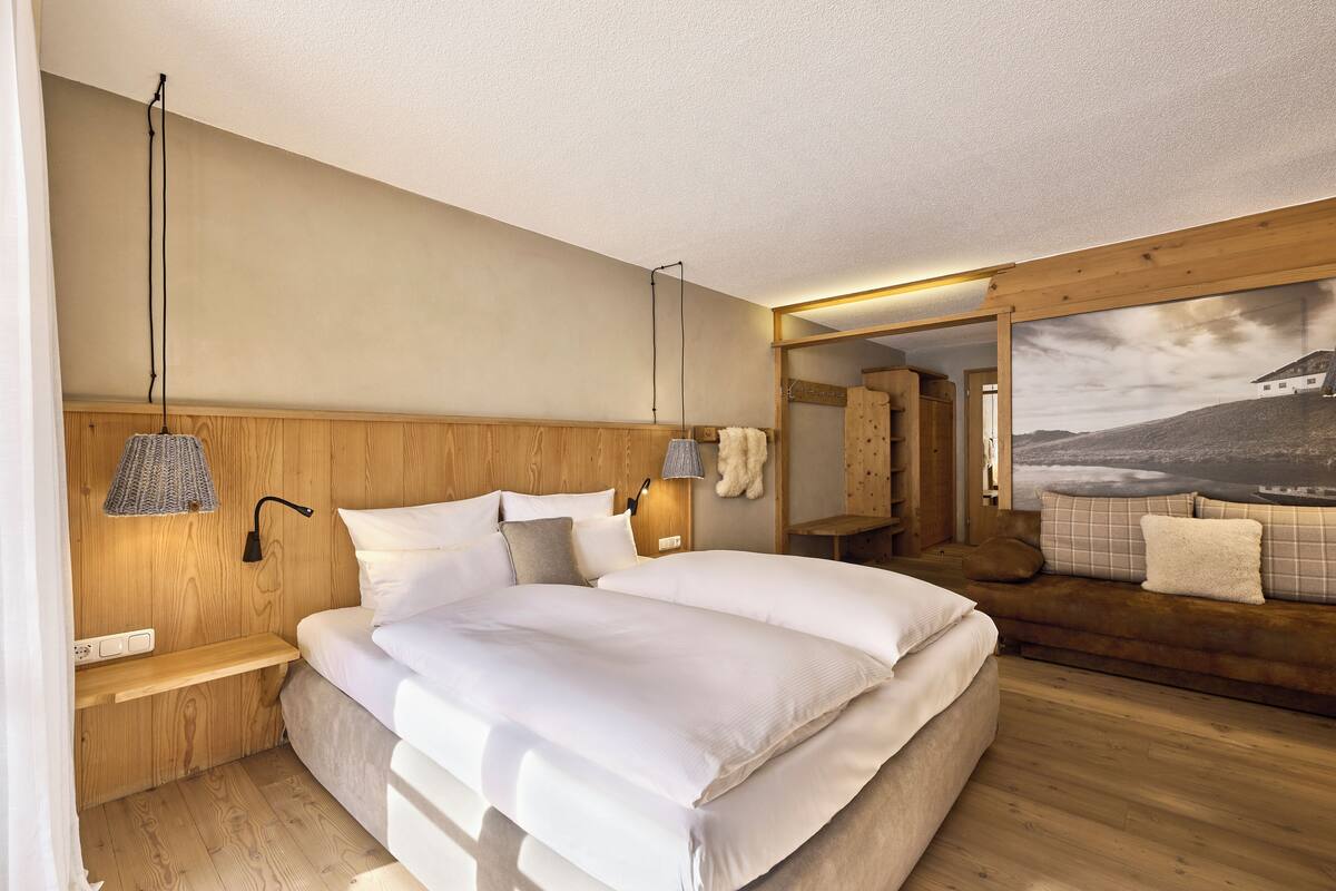 A modern bedroom with a large bed, wooden headboard, bedside tables, a wall-mounted lamp, and a large window.