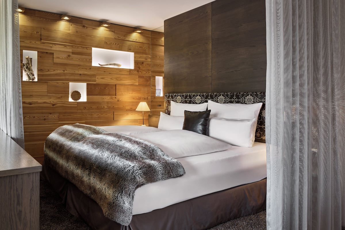 A modern hotel room with a wooden wall panel design, a large bed with white linens, a fluffy throw blanket, and a bedside table with a lamp.