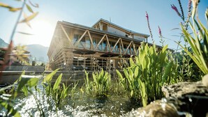 A modern building with a glass facade and a deck extending over a body of water, surrounded by greenery. - Hubertus Mountain Refugio Allgäu (Balderschwang)