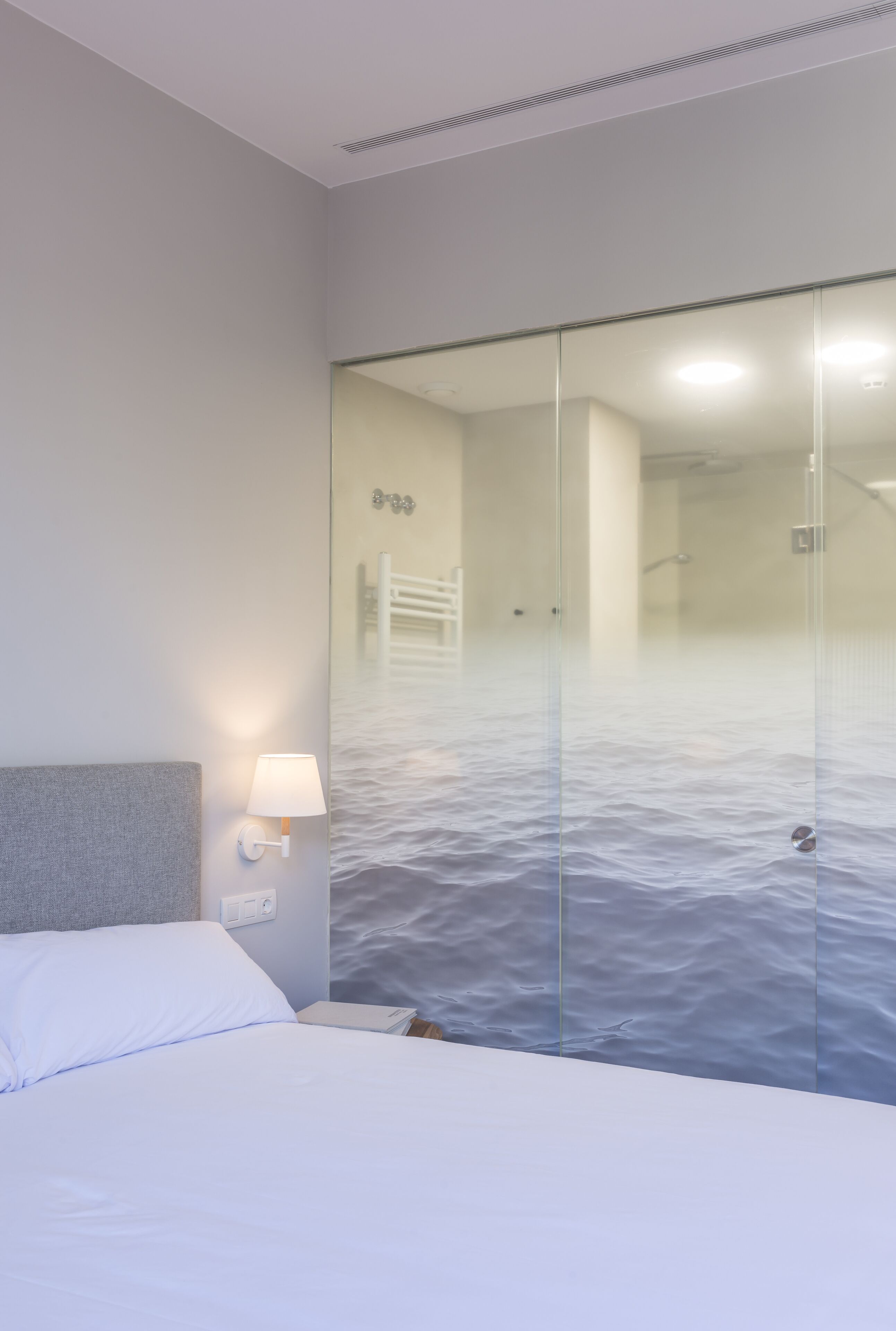 Camera d'albergo con un letto, un comodino, una lampada a parete e una porta in vetro che dà su un bagno con doccia.