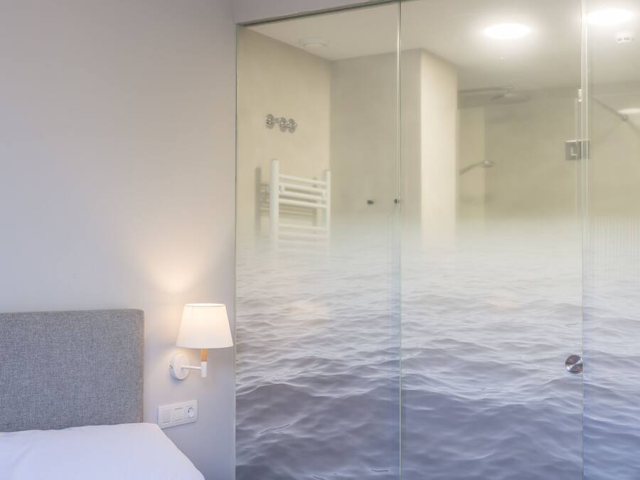 Camera d'albergo con un letto, un comodino, una lampada a parete e una porta in vetro che dà su un bagno con doccia.