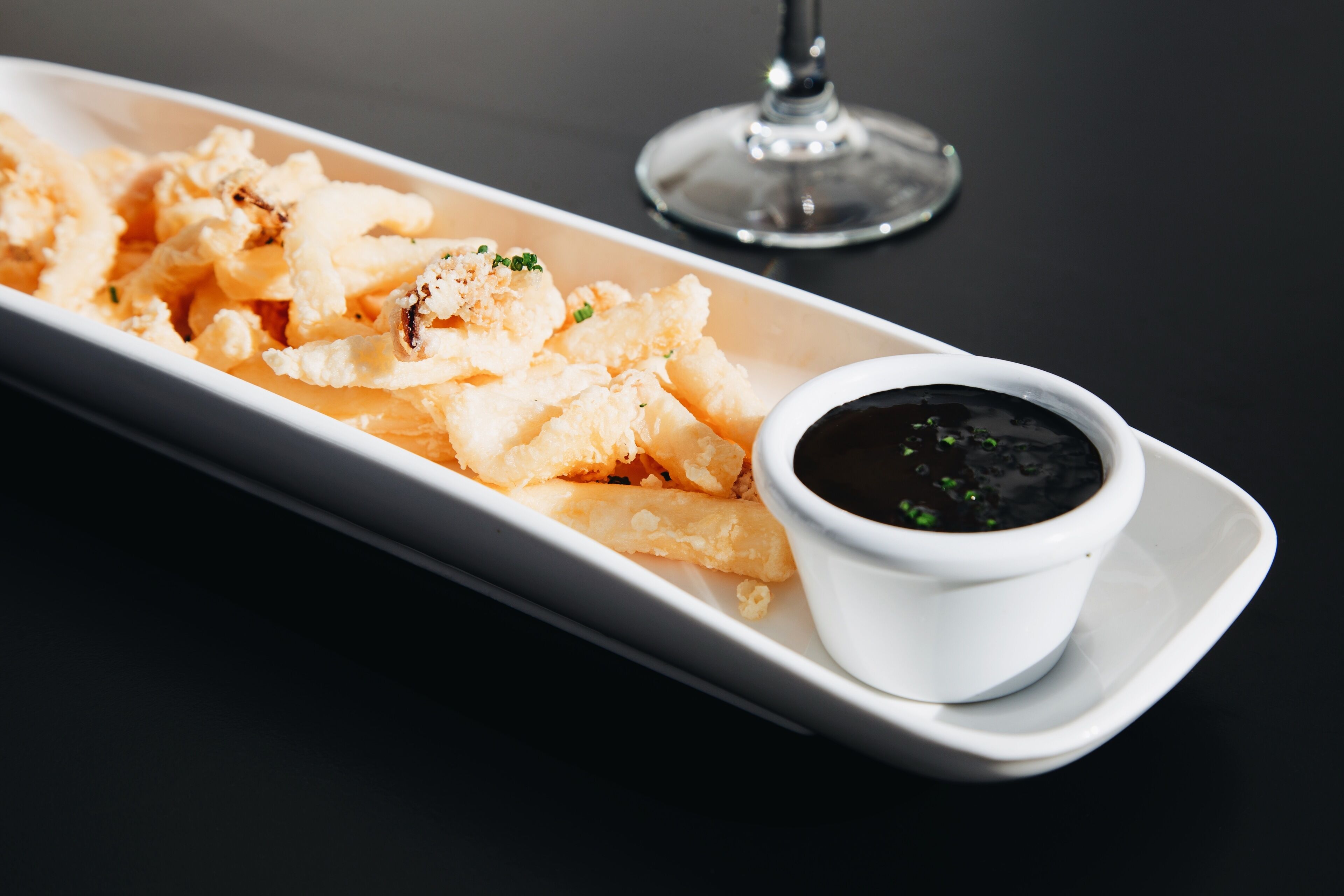 A white rectangular plate with fried calamari and a small bowl of black dipping sauce on a dark table.