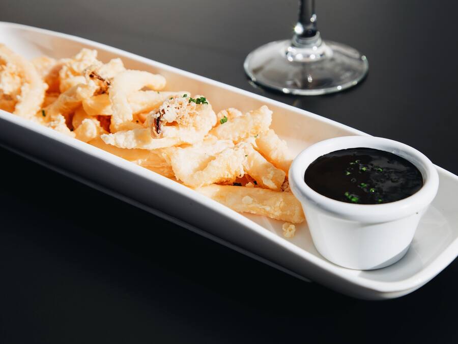 A white rectangular plate with fried calamari and a small bowl of black dipping sauce on a dark table.