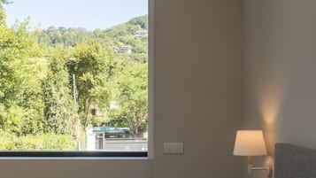 A bedroom with a bed, a window with a view of greenery and buildings, and a bedside lamp.