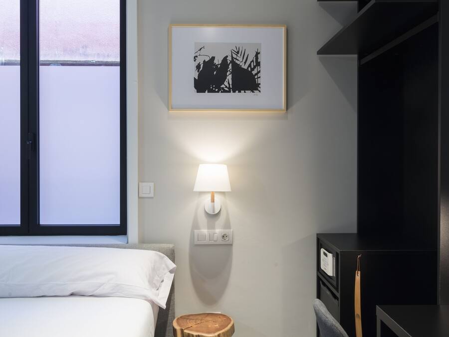 A hotel room with a bed, a desk, a chair, a small stool, a wall-mounted lamp, a framed picture, and a shelving unit.