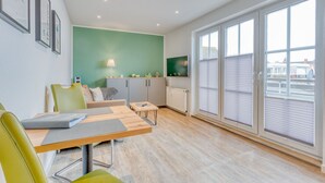 A modern living room with a green accent wall, a dining area with a wooden table, and large windows with blinds.
