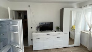 A modern living room with a white entertainment unit, a flat-screen TV, and a large window with sheer curtains. - ZV-Scholtz Gästehaus Neuötting (Neuoetting)