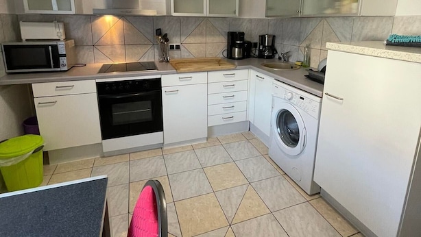 A compact kitchen with white cabinets, a microwave, a stove, a washing machine, and a dining area with a chair. - ZV-Scholtz Gästehaus Neuötting (Neuoetting)