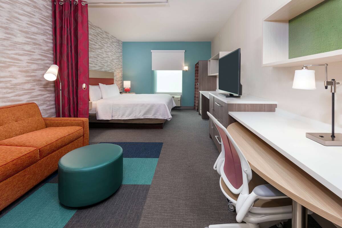 Habitación de hotel con cama, sofá, escritorio con silla, televisor y ventana con cortina.