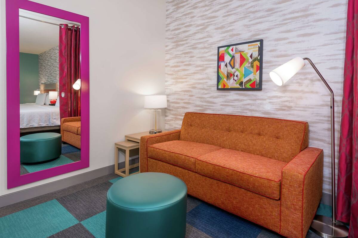 Habitación de hotel con pared espejada, un sofá, un taburete redondo, una lámpara y un cuadro abstracto de colores.