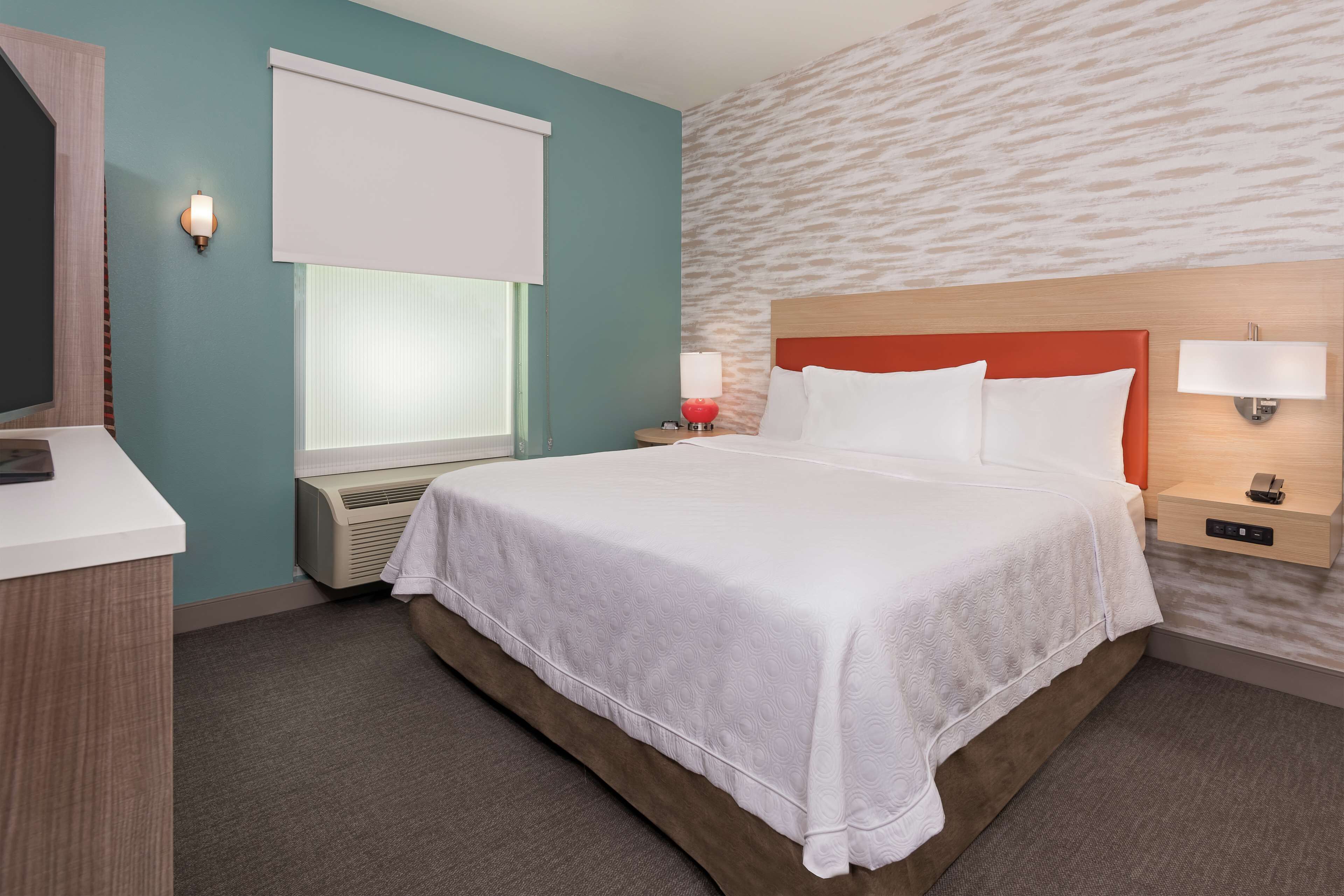 A hotel room with a large bed, a desk, a window with blinds, a wall-mounted lamp, and a thermostat.