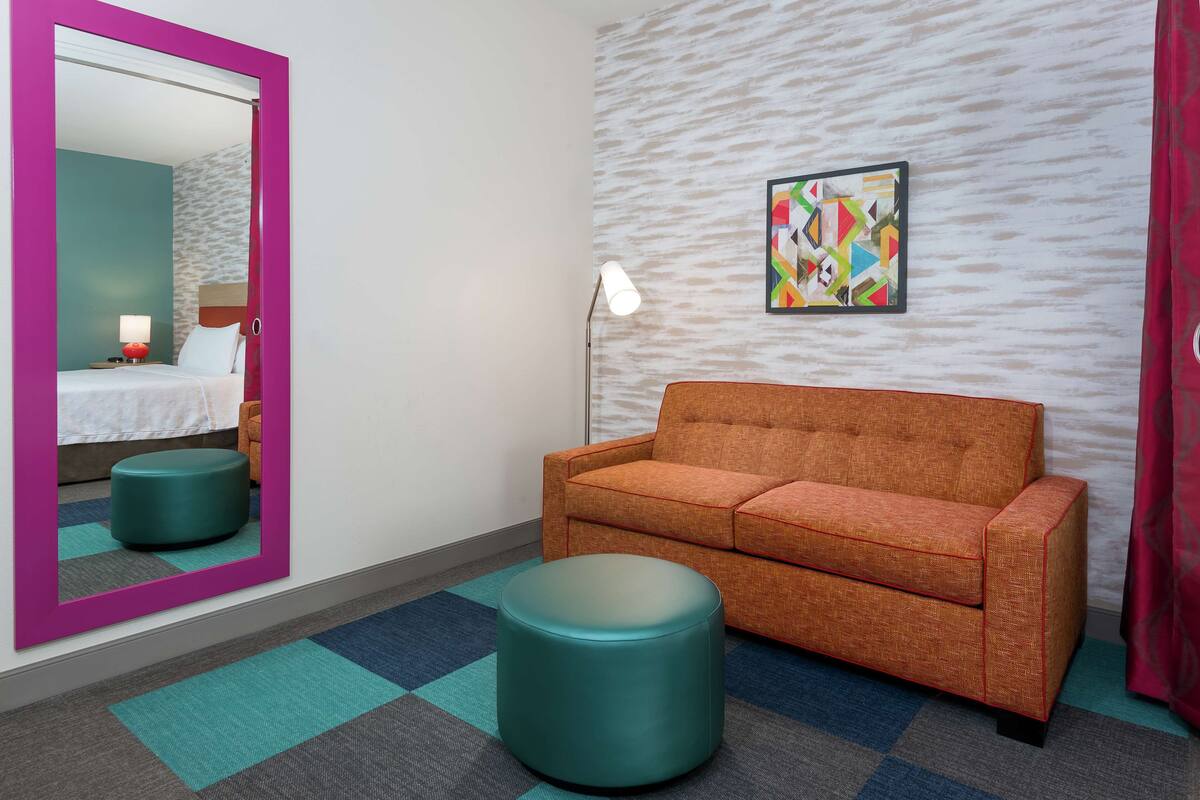 Habitación de hotel con un reposapiés verde azulado, un sofá naranja, un espejo grande con marco rosa y un cuadro abstracto y colorido en la pared.