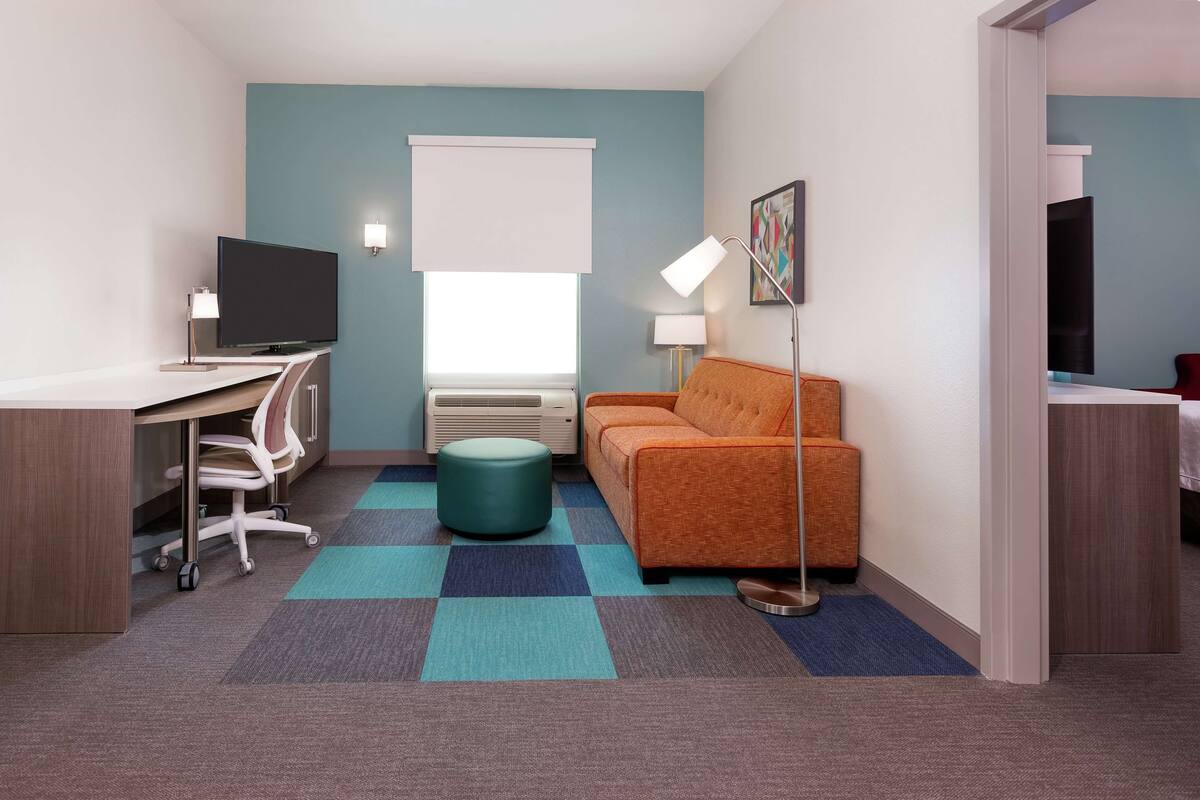 Habitación de hotel compacta con escritorio, silla, televisor, sofá y alfombra estampada.