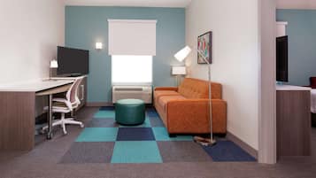 A compact hotel room with a desk, chair, television, sofa, and a patterned rug.