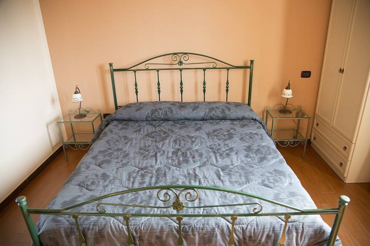 A single bed with a metal frame and a quilted mattress in a room with wooden flooring and peach-colored walls.