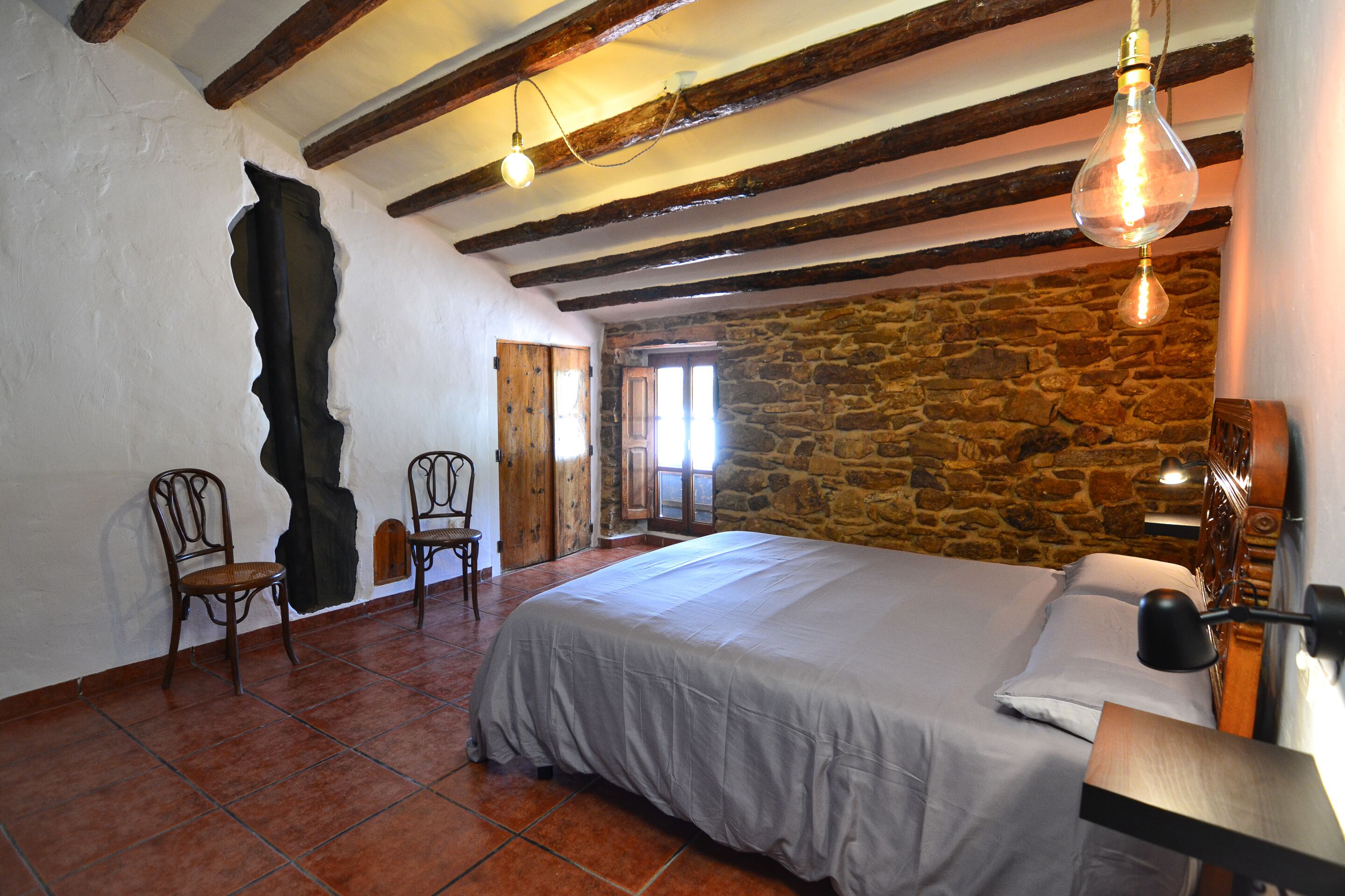 Une chambre avec un mur en pierre, des poutres en bois, un lit recouvert d’une couverture grise, deux chaises et un luminaire suspendu.