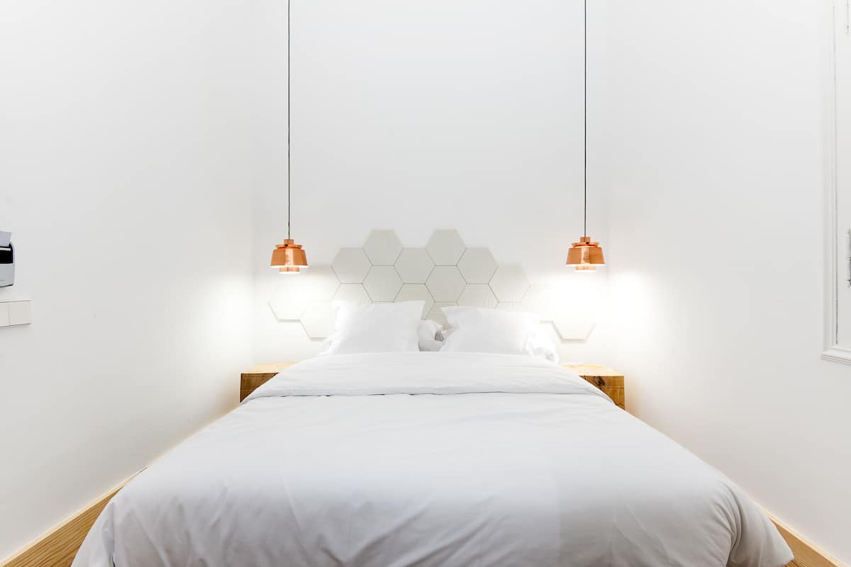 A minimalist bedroom with a white bed, wooden headboard, and two pendant lights.