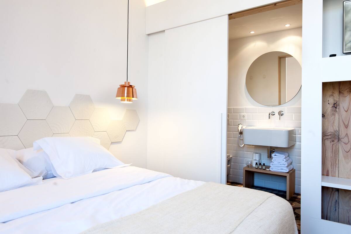 A modern hotel room with a neatly made bed, a round mirror above the sink, and a hexagonal wall design.