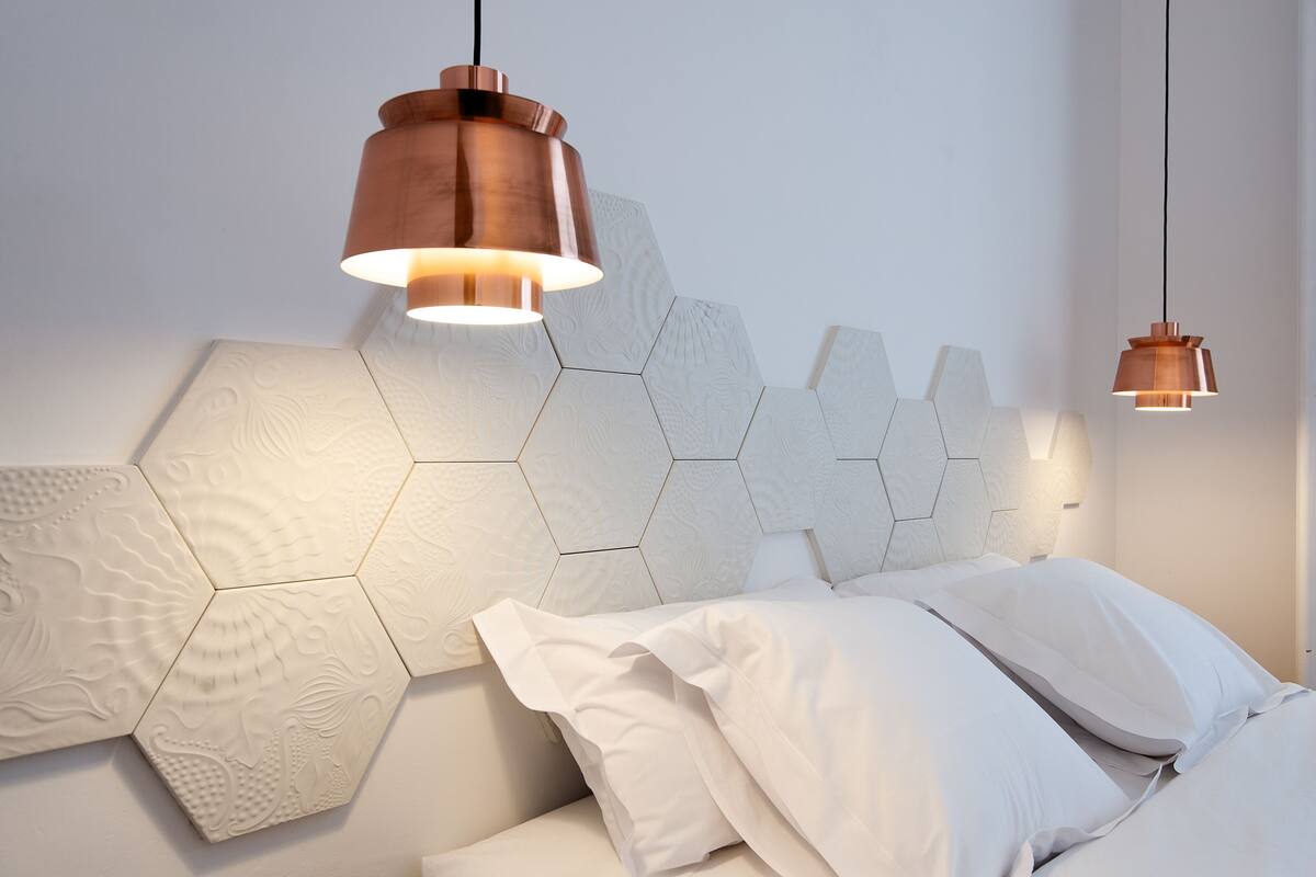 A modern bedroom with a hexagonal patterned headboard, a white bed with multiple pillows, and two copper pendant lights.