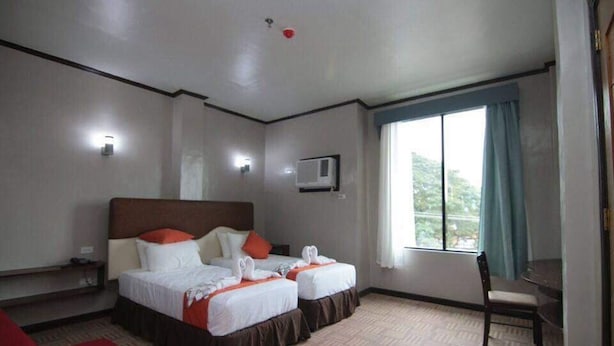 A hotel room with two beds, a desk, a chair, a window with curtains, and an air conditioning unit.