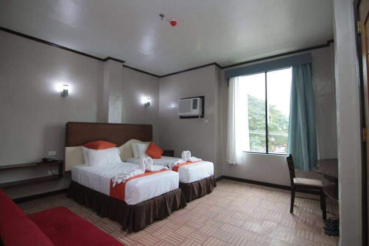 A hotel room with two beds, a desk, a chair, a window with curtains, and an air conditioning unit.