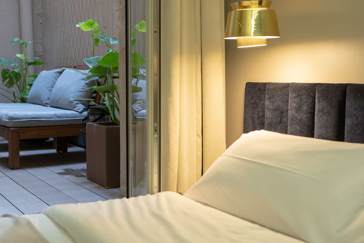 Een slaapkamer met een bed, nachtkastje, lamp, raam en uitzicht op de natuur.
