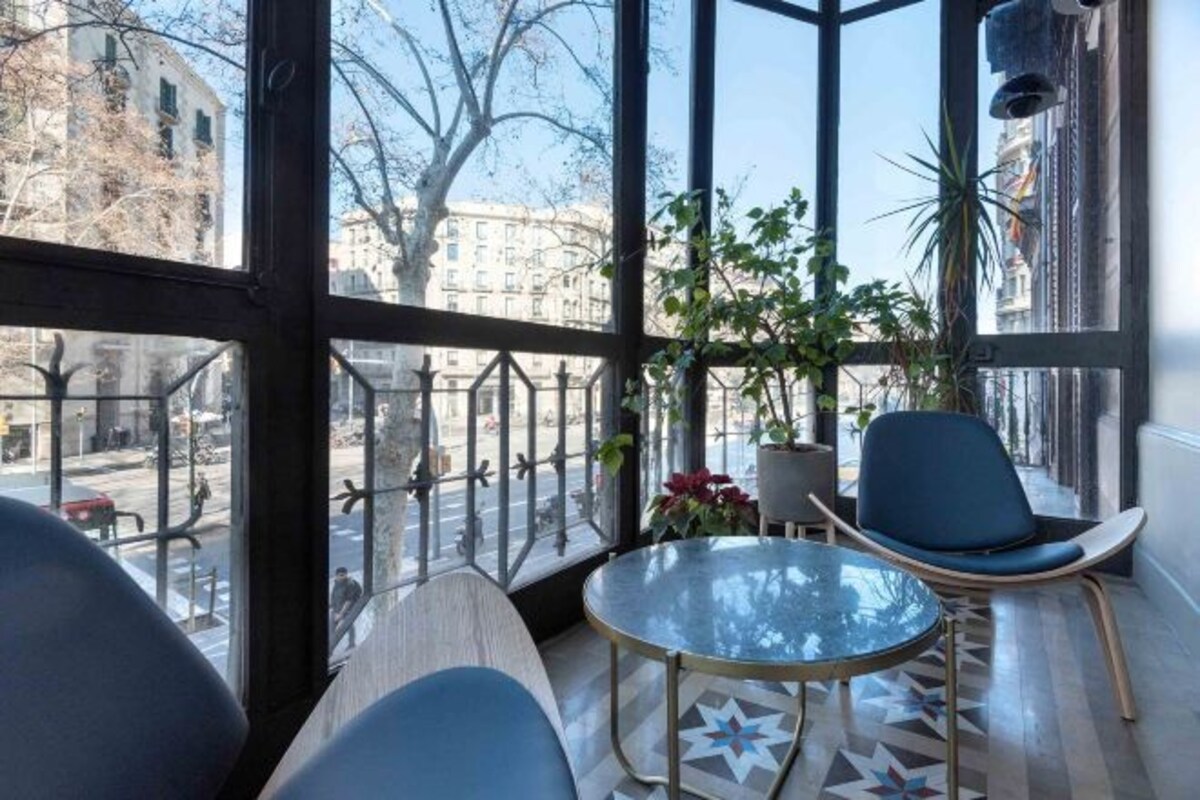 A modern room with large windows, a round glass table, two blue chairs, and a potted plant.