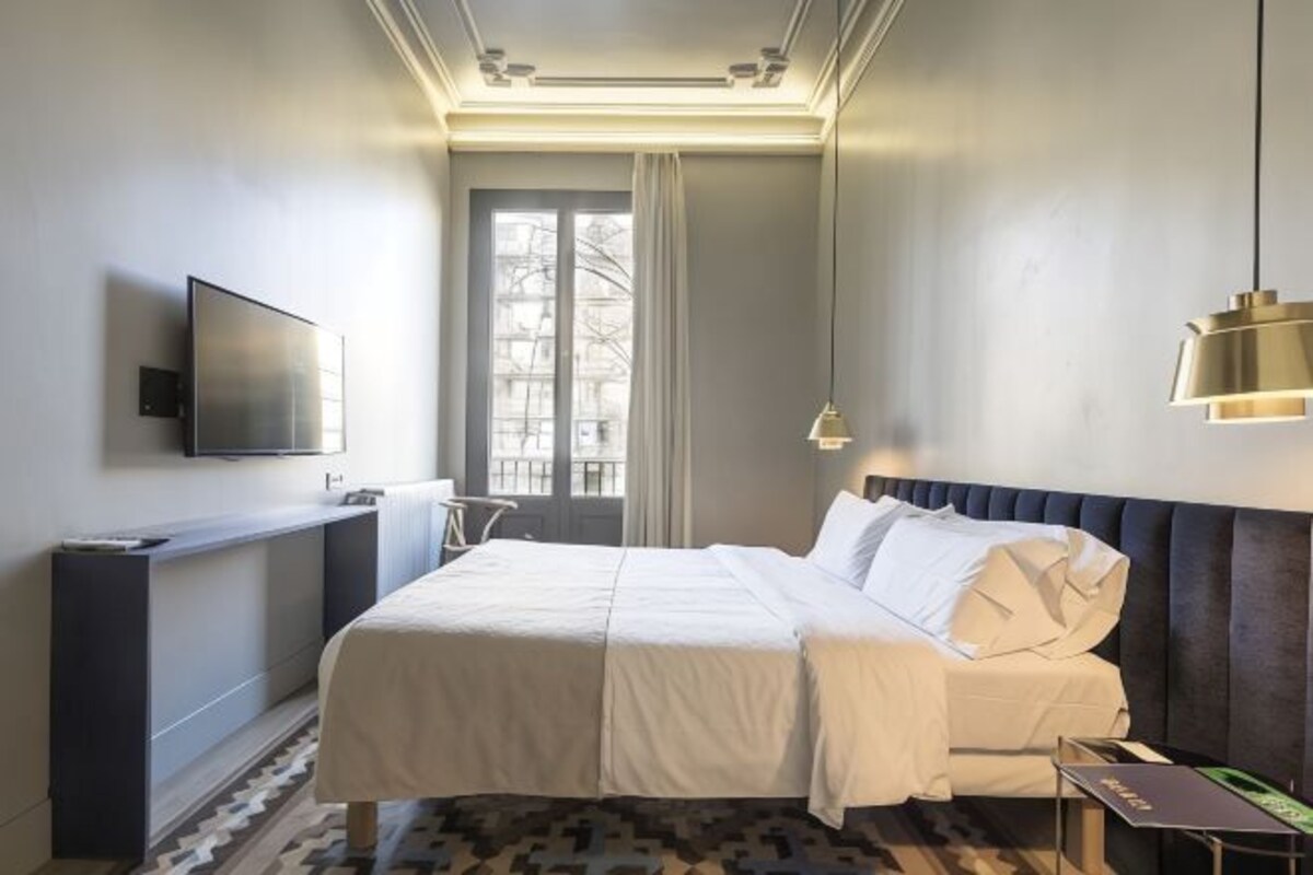 A modern hotel room with a large bed, a flat-screen TV mounted on the wall, a desk, and a window with curtains.