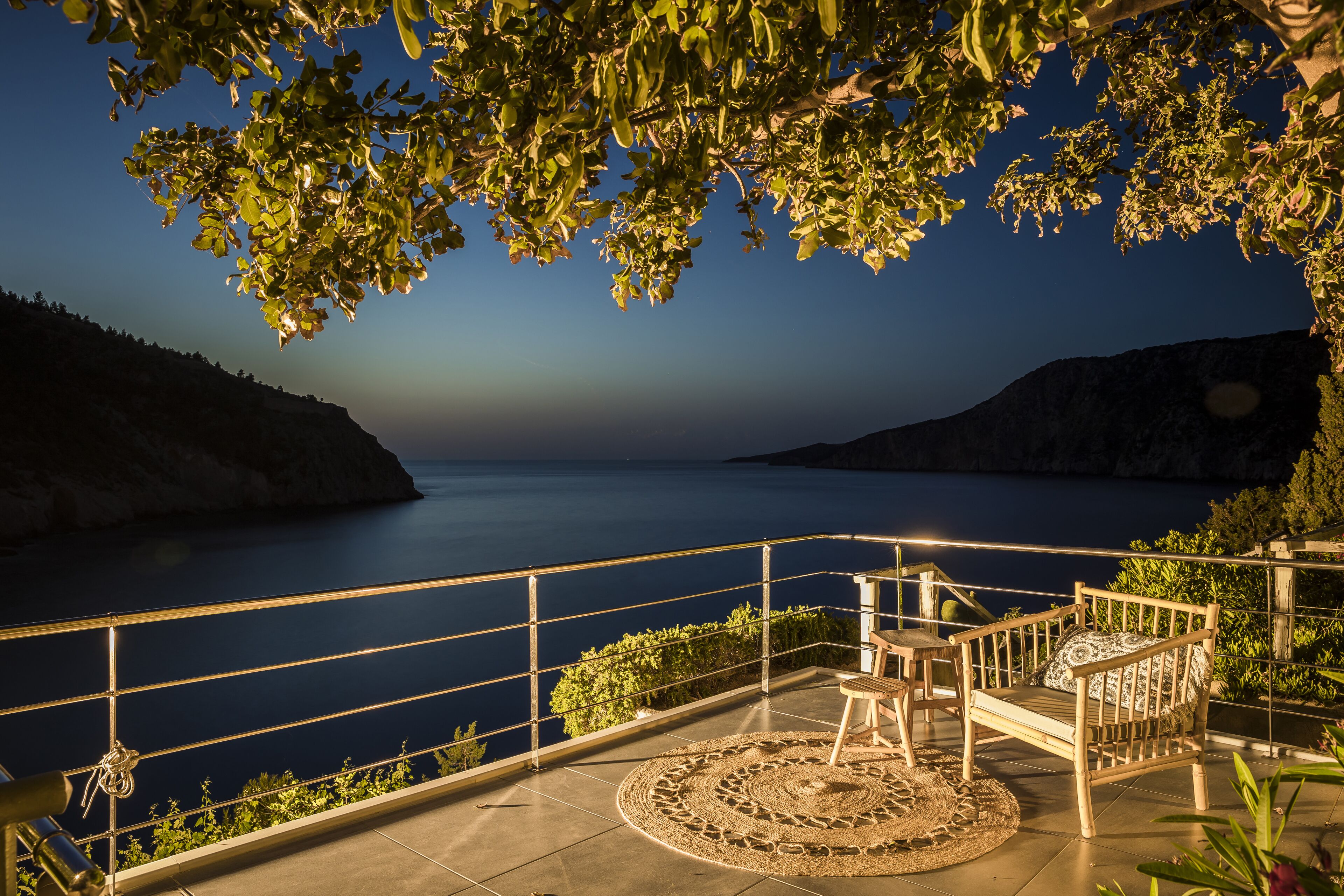 A balcony with a view of the sea, a wooden chair, and a table with a patterned rug.