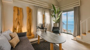 A modern living room with a sofa, a coffee table, and a large plant. There is a view of the ocean through the windows.