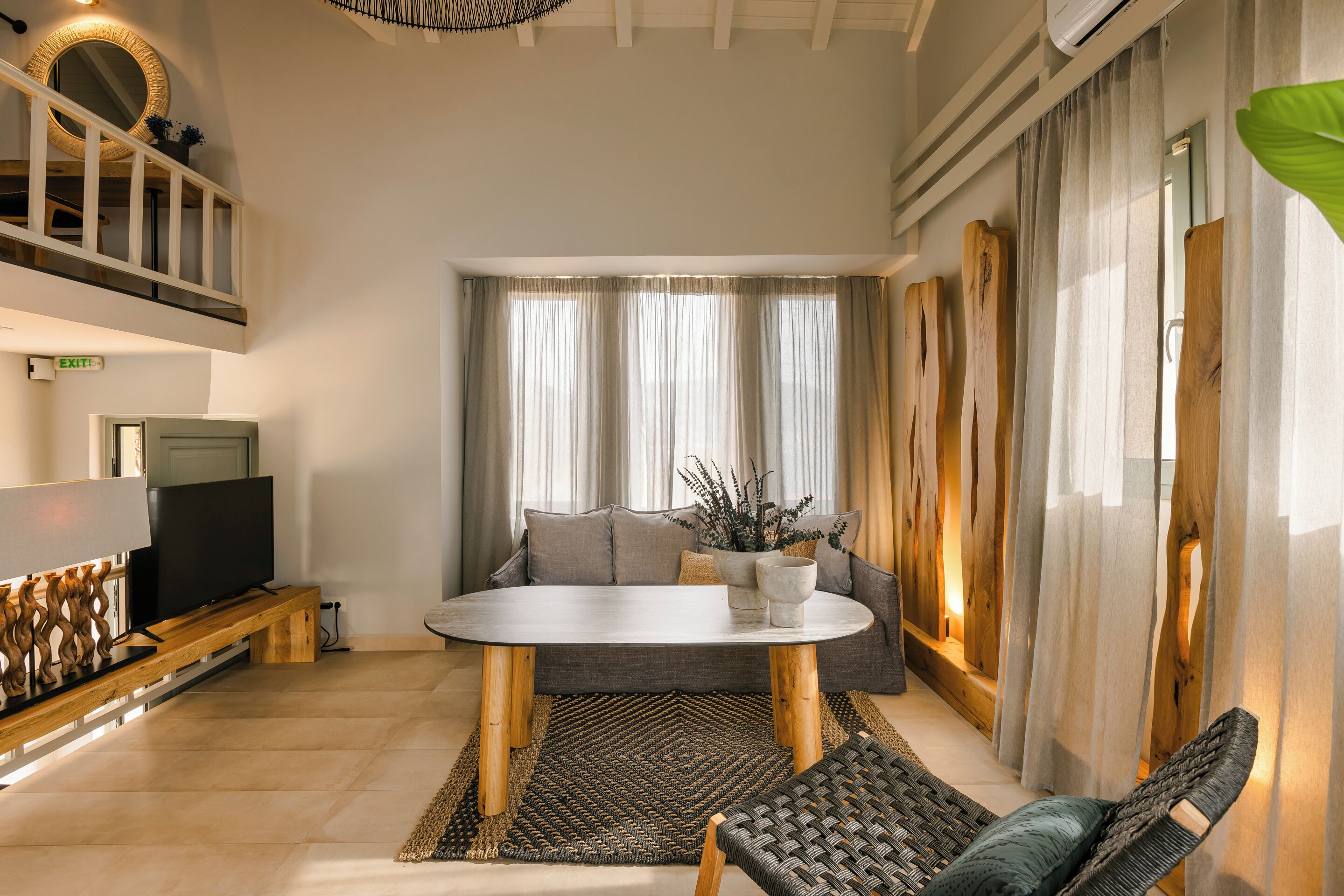 Een moderne woonkamer met een ronde tafel, een bank en twee stoelen. Er is een televisie op een houten dressoir, een groot raam met gordijnen en een plafondventilator.