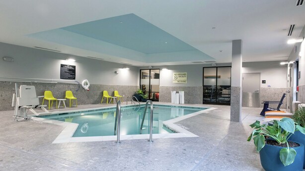 An indoor swimming pool with clear water, surrounded by grey marble flooring. There are yellow chairs and a blue potted plant near the pool. - Hampton Inn Black Mountain (Black Mountain)