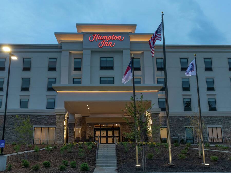 A Hampton Inn hotel with a modern facade, a well-lit entrance, and American flags.