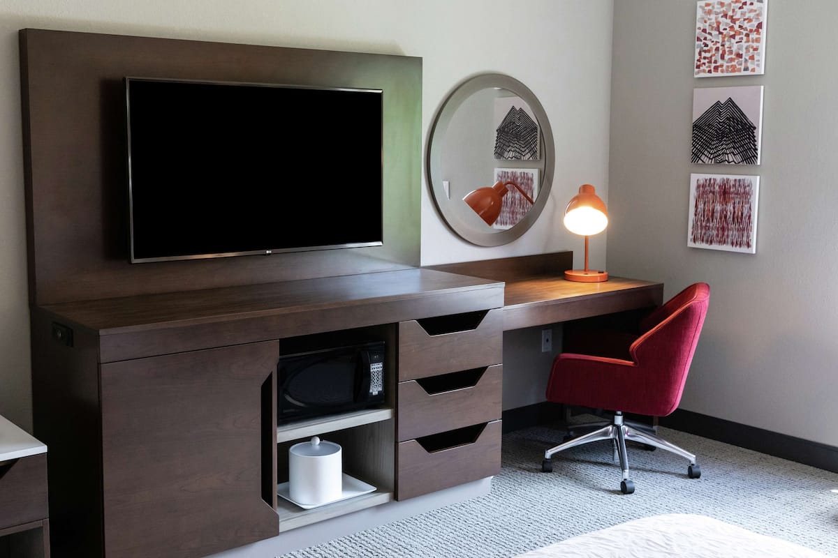 A modern hotel room with a large flat-screen TV mounted on a dark wood cabinet, a red office chair, a round mirror, and three framed artworks on the wall.