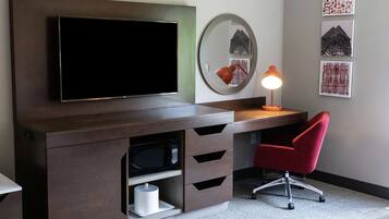 Una habitación de hotel moderna con un televisor de pantalla plana grande montado en un mueble de madera oscura, una silla de escritorio roja, un espejo redondo y tres obras de arte enmarcadas en la pared.