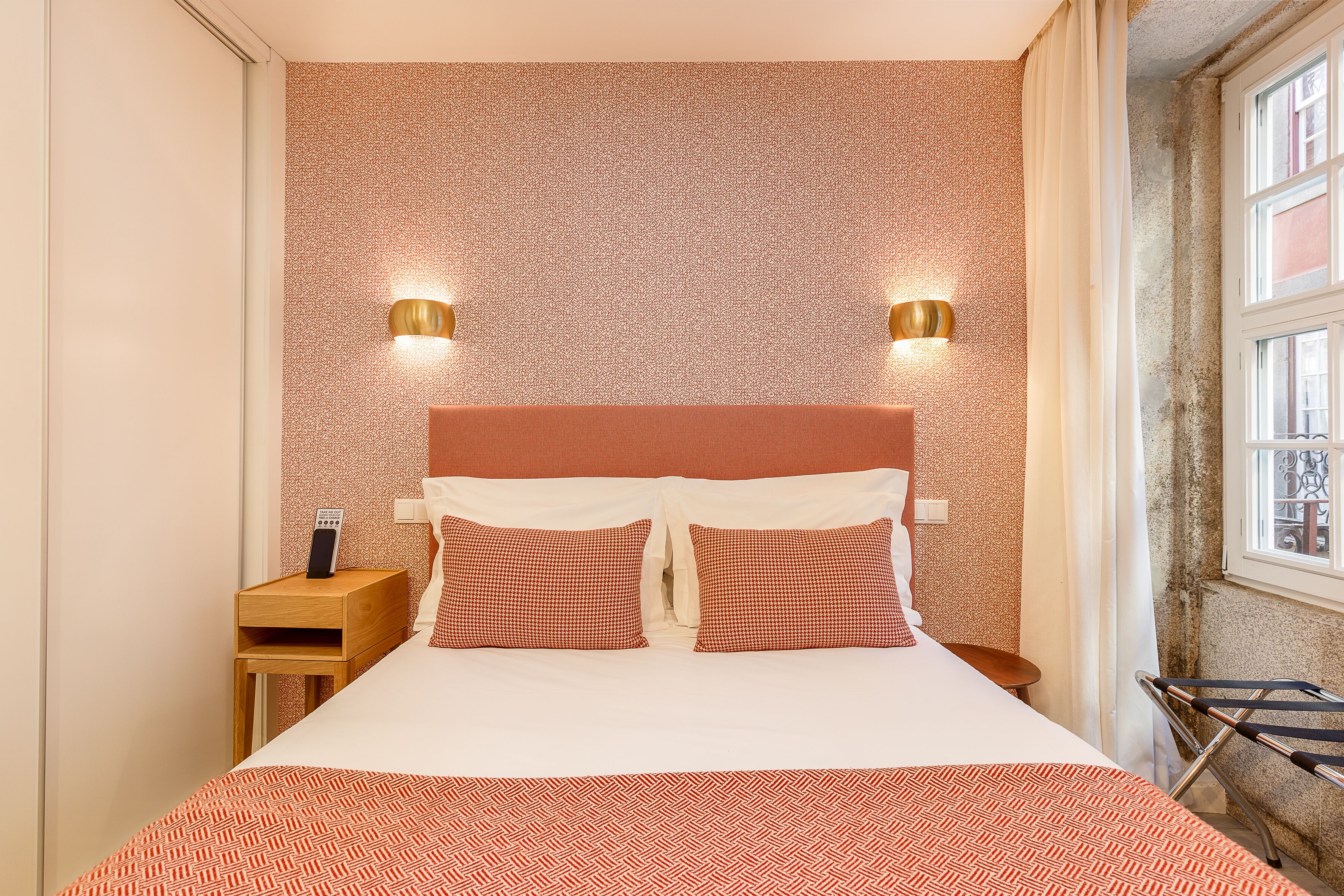A neatly made bed with a patterned bedspread, two pillows, and a headboard. There are two wall-mounted lights and a small wooden nightstand with a phone.