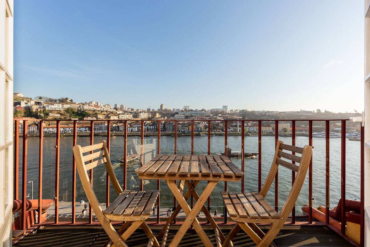 A balcony with a wooden table and chairs, overlooking a waterfront with buildings and boats.