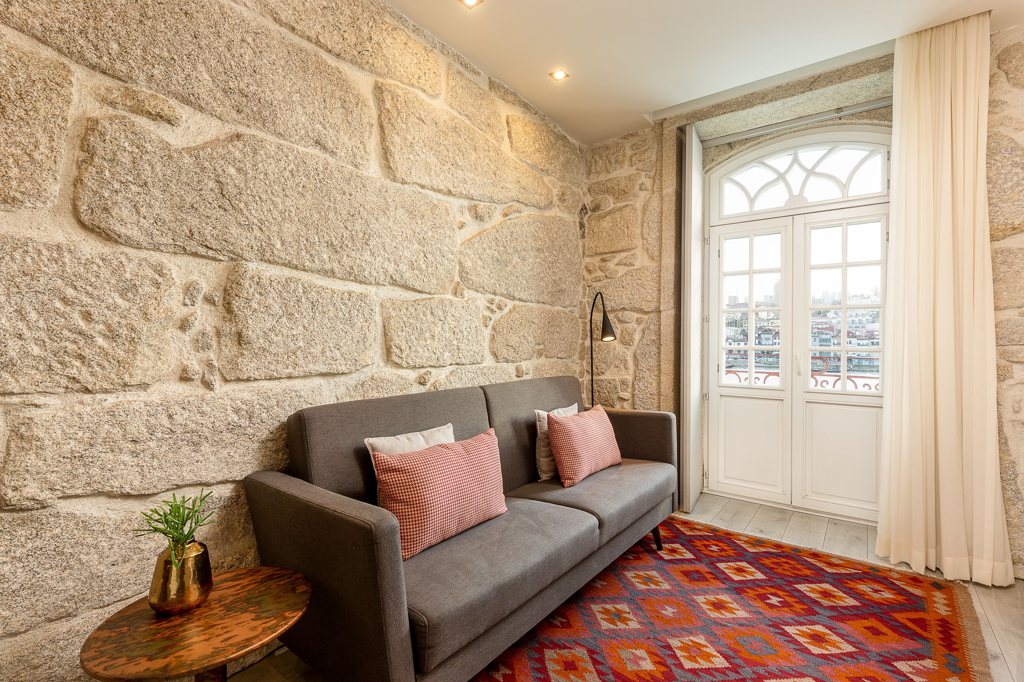 A room with a stone wall, a grey sofa with red and white cushions, a small round table with a plant, and a colorful rug.