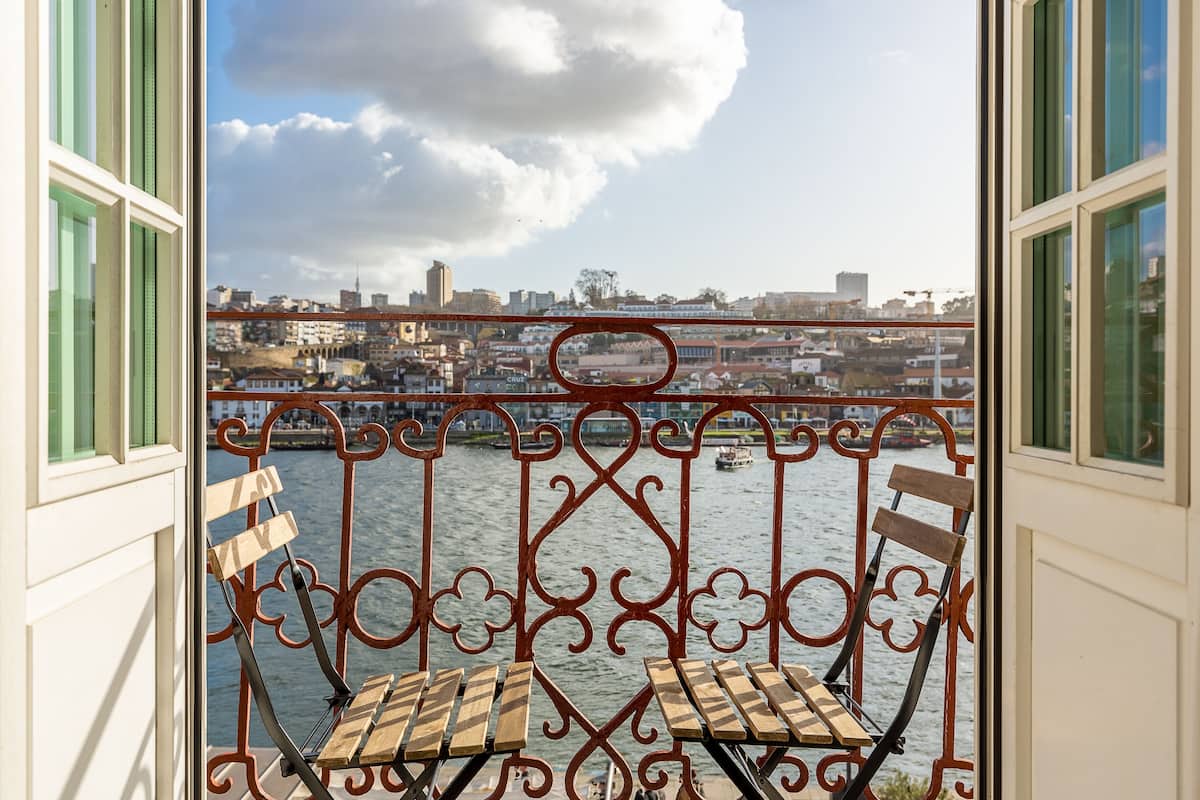 A balcony with a wrought iron railing and two wooden chairs, overlooking a river with buildings in the background.